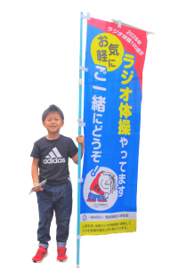 ラジオ体操やってますお気軽にどうぞと書いてある旗を持つ凌平君